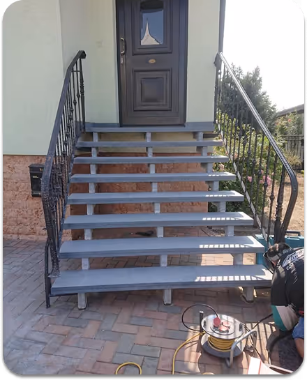 Außentreppe mit grauen Stufen und schwarzen Metallgeländern, führt zu einer dunklen Haustür, Handwerker arbeitet unten rechts.